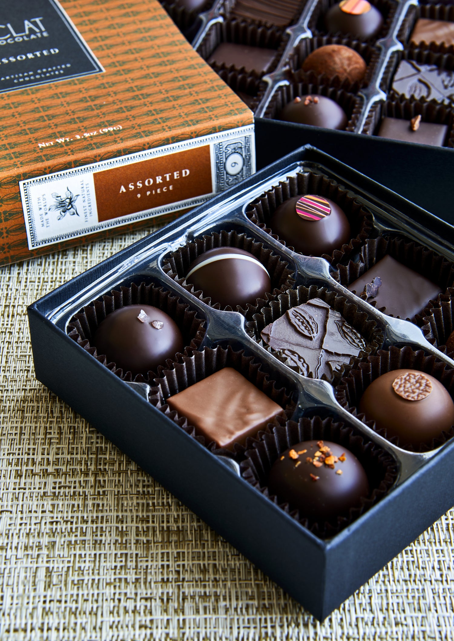 An open black box of assorted chocolate truffles and squares with their branded box lid and outer box visible behind it on a woven beige surface.