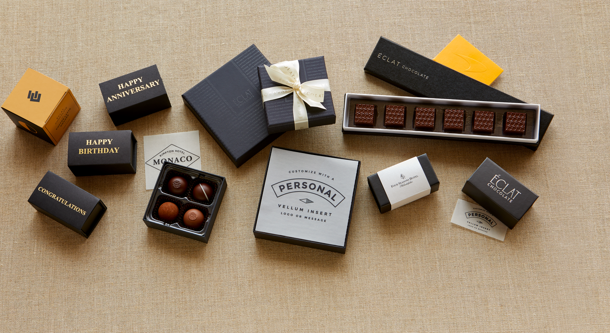 An assortment of premium chocolate gift boxes and open boxes of chocolates arranged on a beige linen background. The boxes are in black, navy, and yellow, with some featuring custom printed text such as 'Happy Birthday', 'Happy Anniversary', and 'Congratulations'.